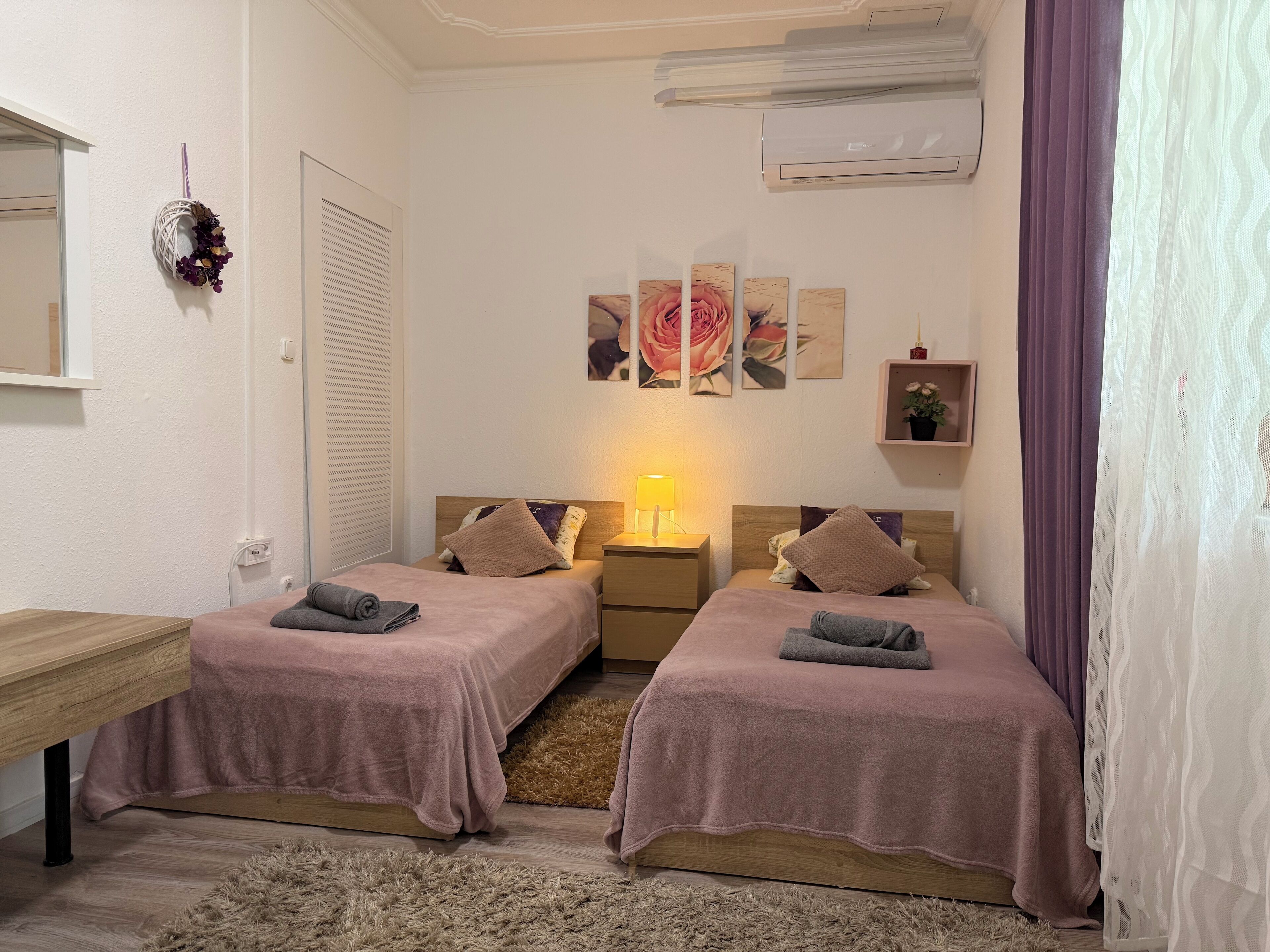 Une chambre avec deux lits, une table de chevet avec une lampe, un climatiseur fixé au mur et une fenêtre avec des stores.