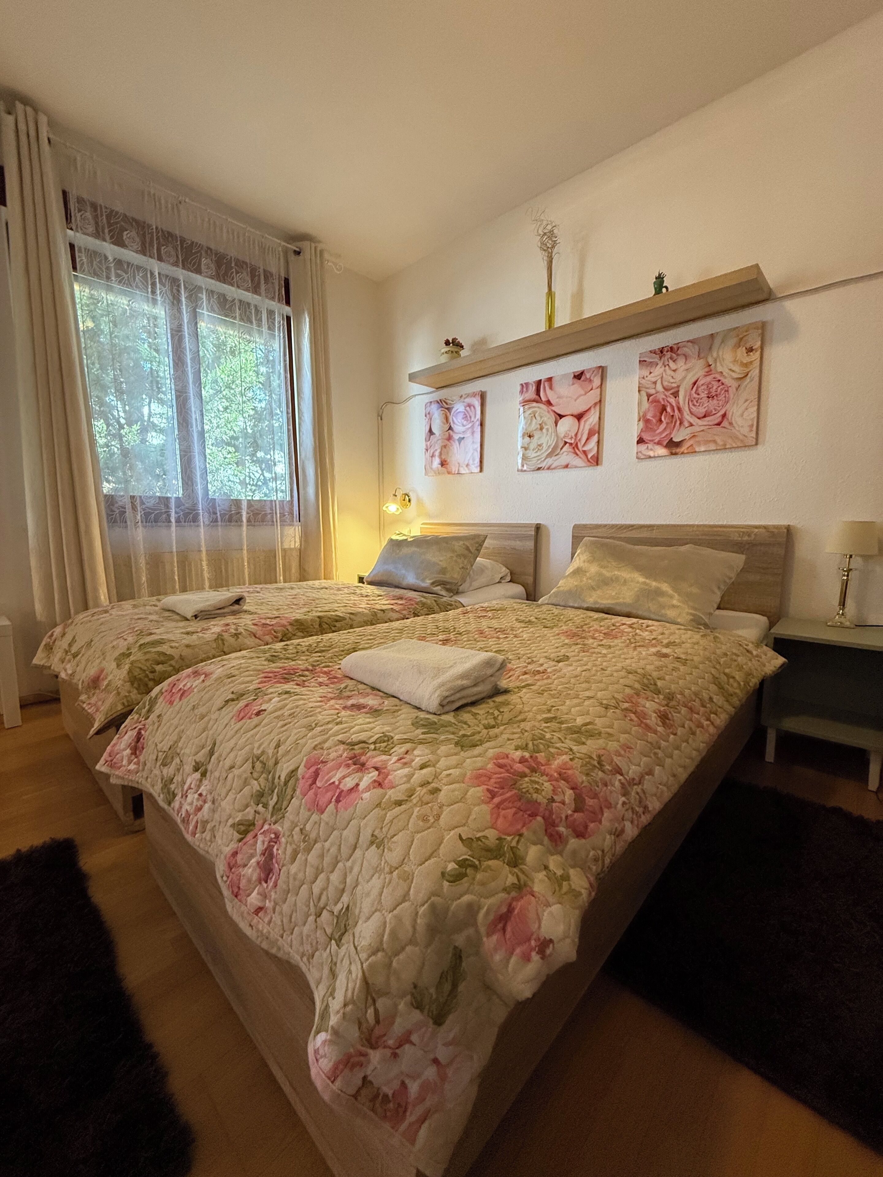 Una cama doble con ropa de cama floral, una mesita de noche y una ventana con cortinas translúcidas.