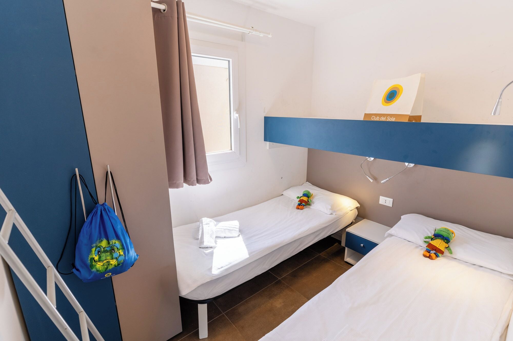 A hotel room with two single beds, a window with curtains, a bedside table with a lamp, and a wall-mounted artwork.