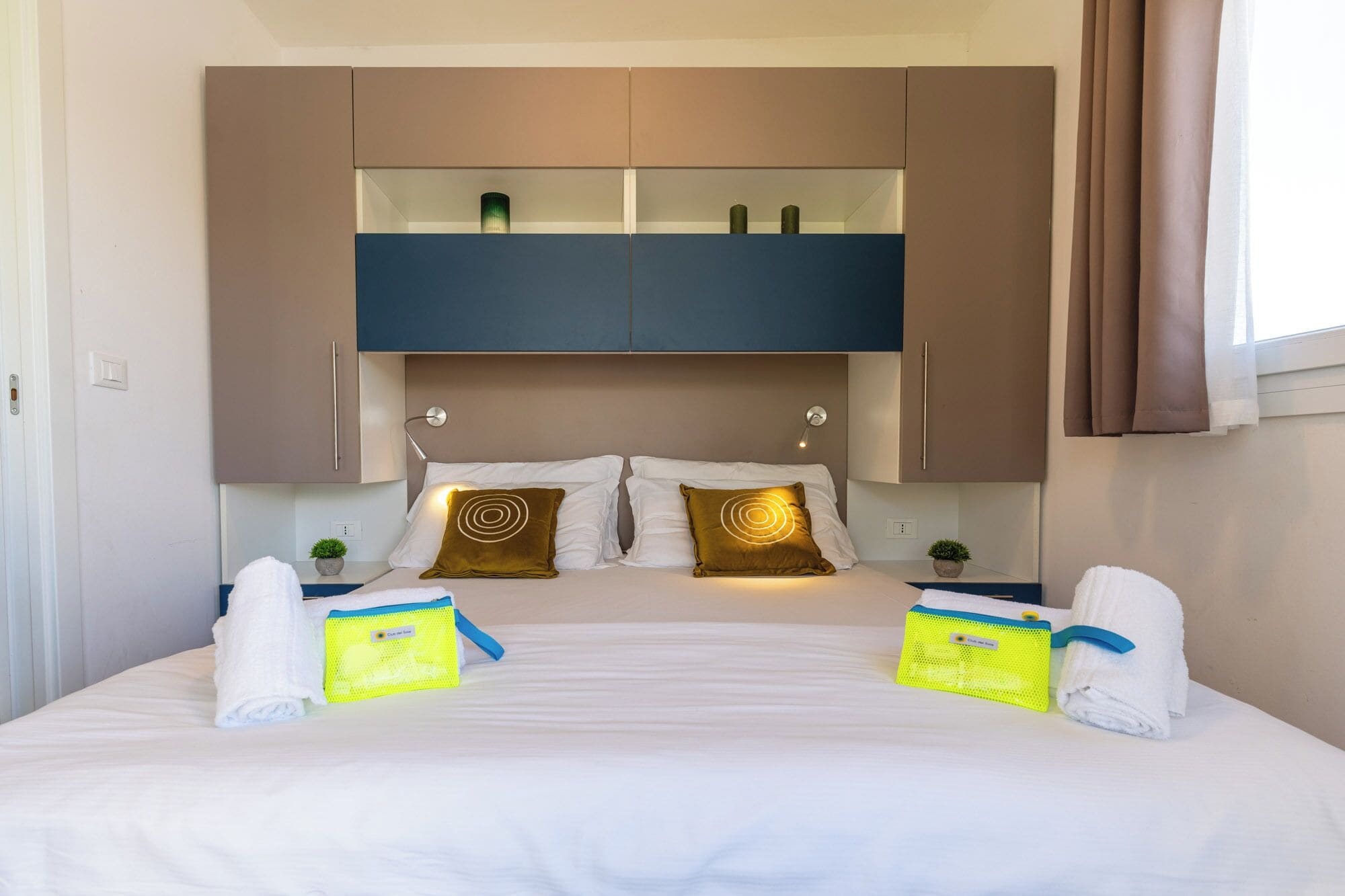 A hotel room with a bed, two bedside tables, a built-in shelf above the bed, and a window with curtains.