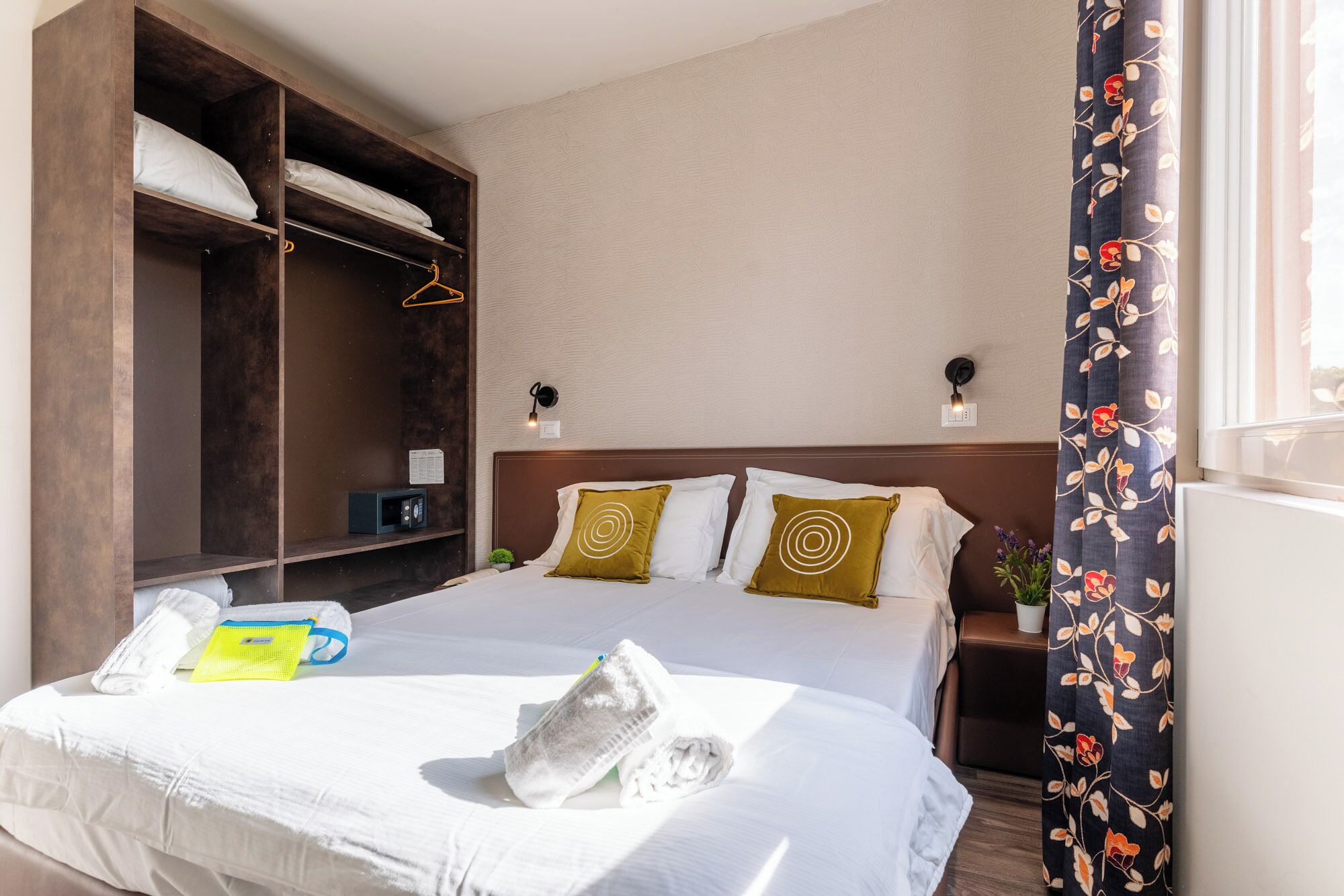 A hotel room with a bed, a wardrobe, a nightstand, a lamp, and a window with curtains.
