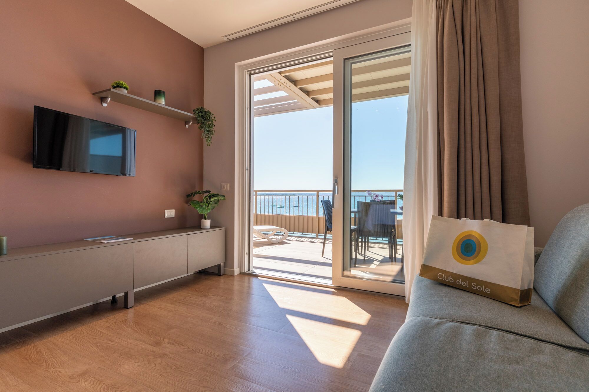A modern living room with a flat-screen TV, a sofa, a dining area with a table and chairs, a balcony with a view of the sea, and a sliding glass door.