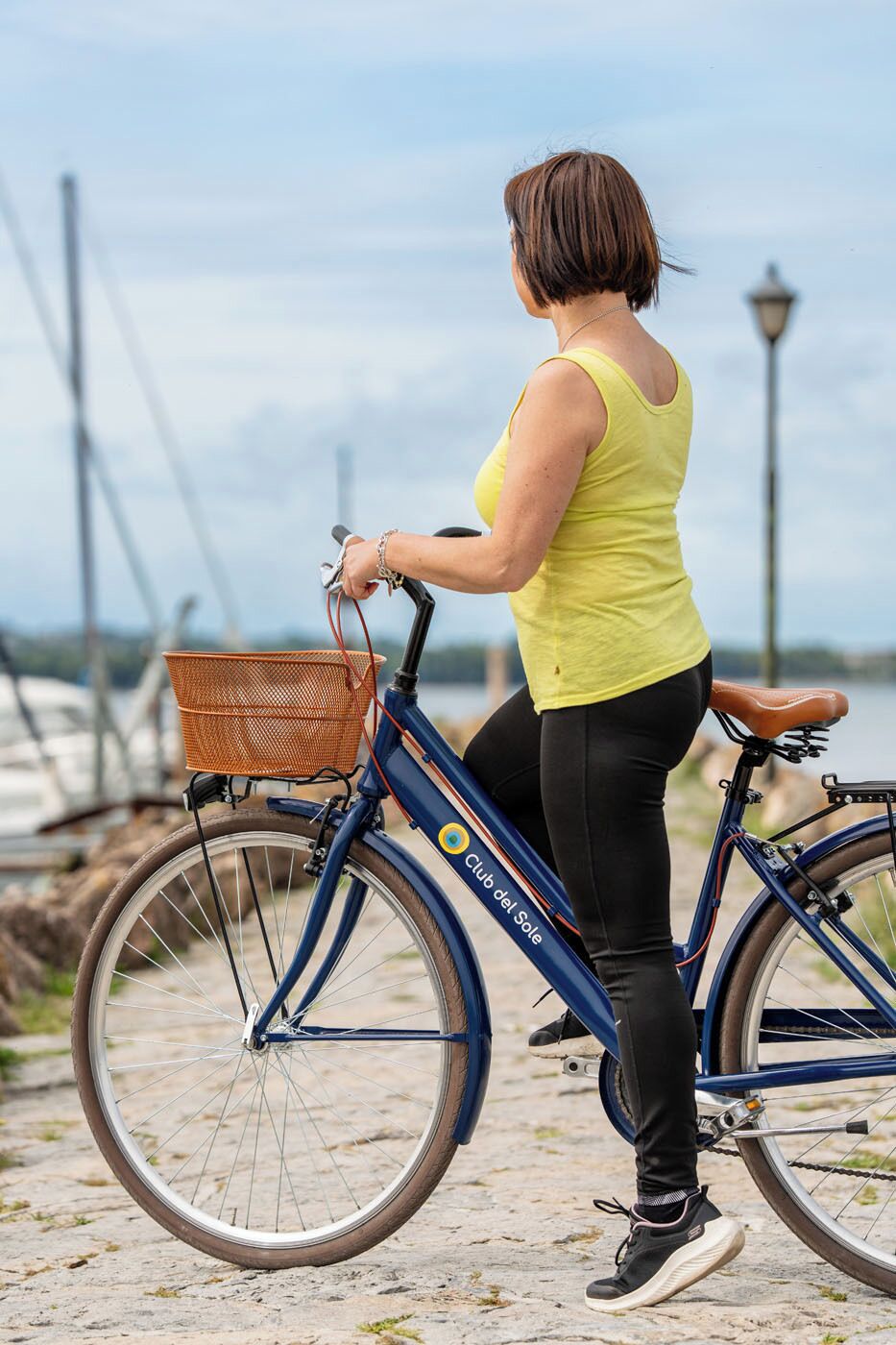 Una persona montando una bicicleta azul con canasta, vestida con una camiseta amarilla y pantalones negros, sobre un camino pavimentado.