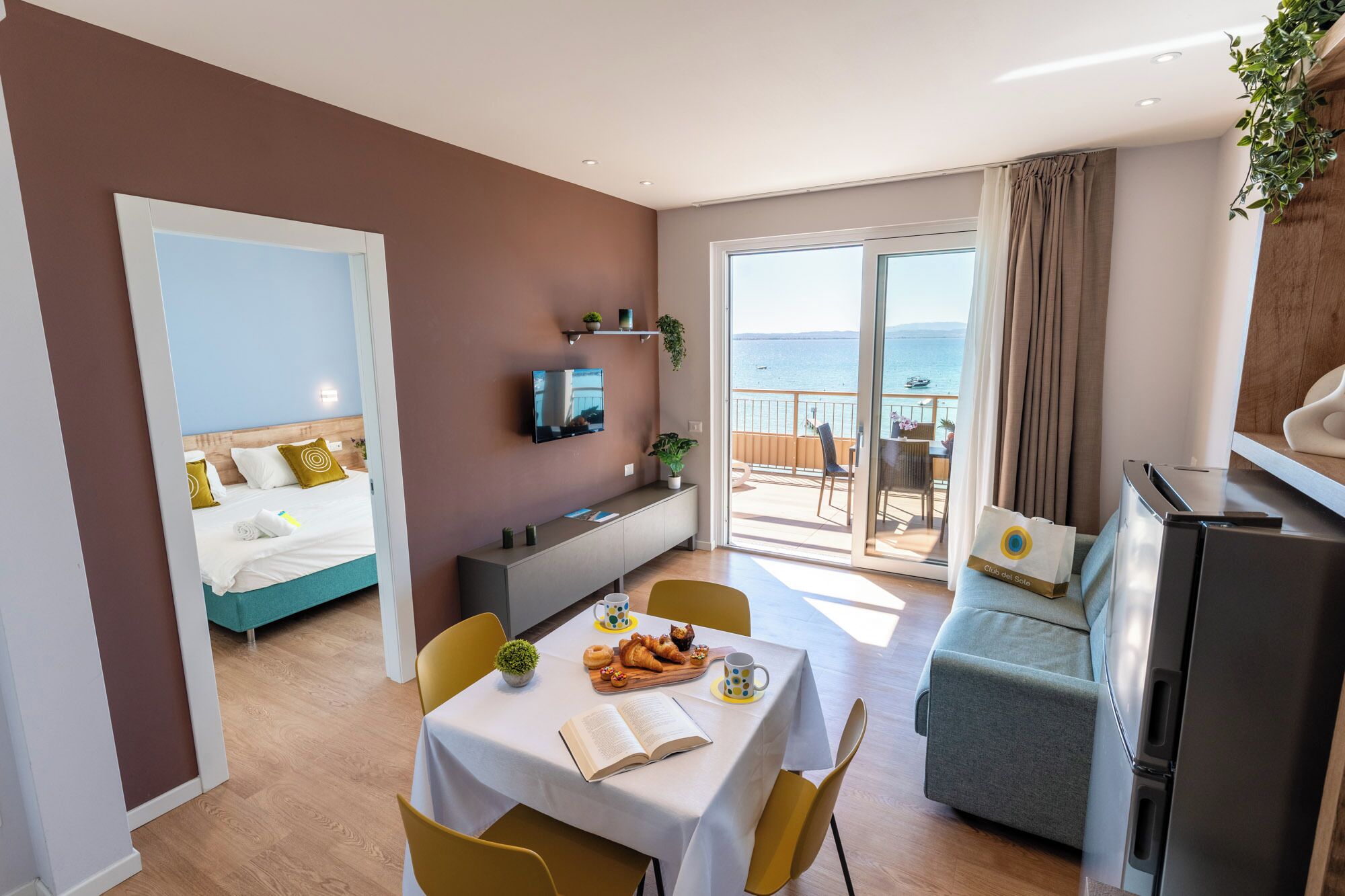 Quarto de hotel com cama, área de jantar com mesa e cadeiras, televisão, varanda com vista para o mar e cozinha compacta.