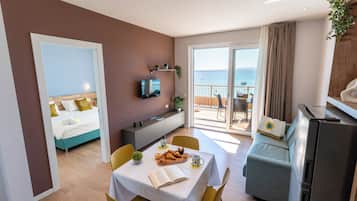 A hotel room with a bed, a dining area with a table and chairs, a TV, a balcony with a view of the sea, and a kitchenette.