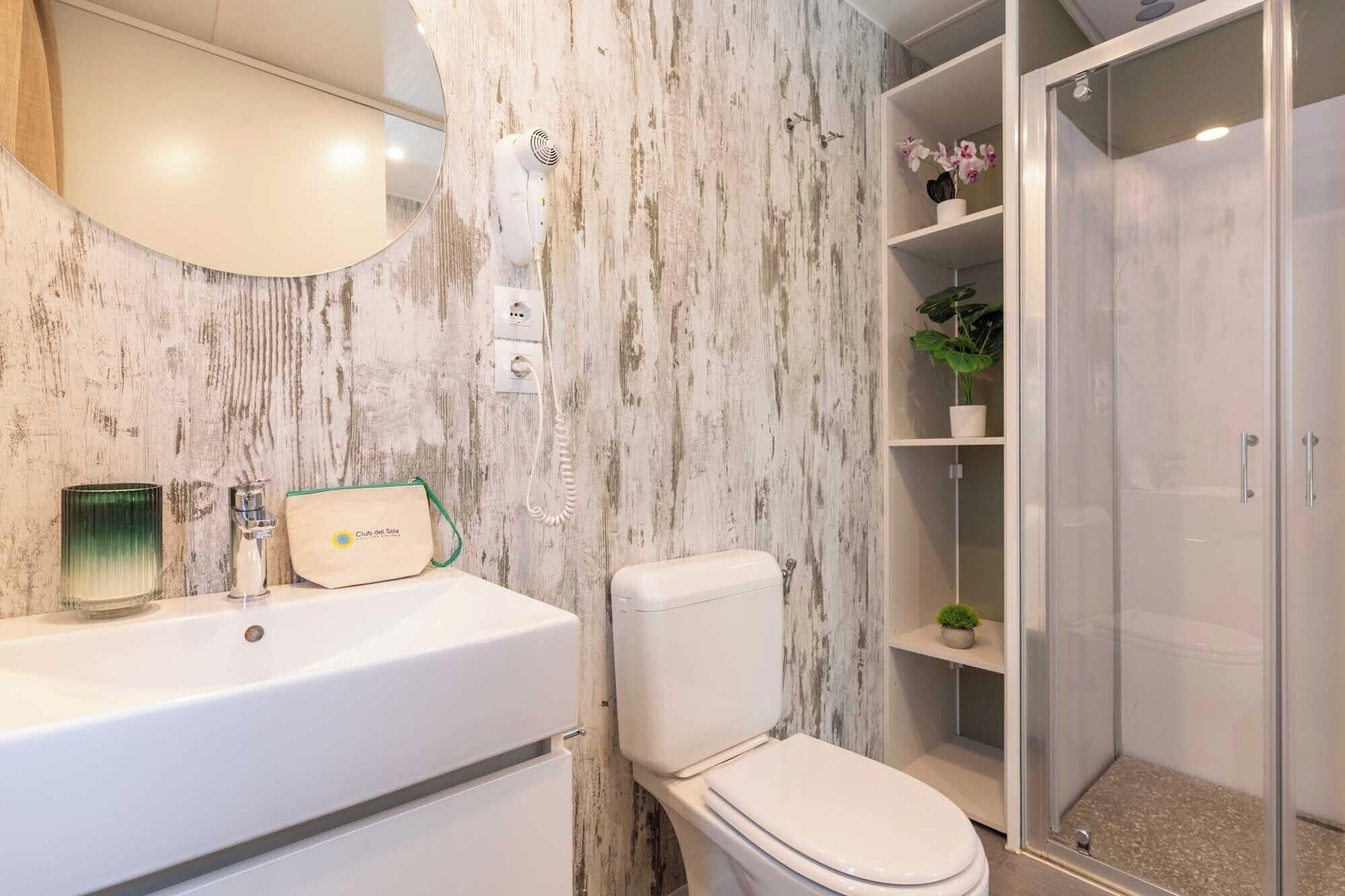 Bagno con parete in marmo, un water bianco, un lavandino con lampada a sfumatura verde e cabina doccia.