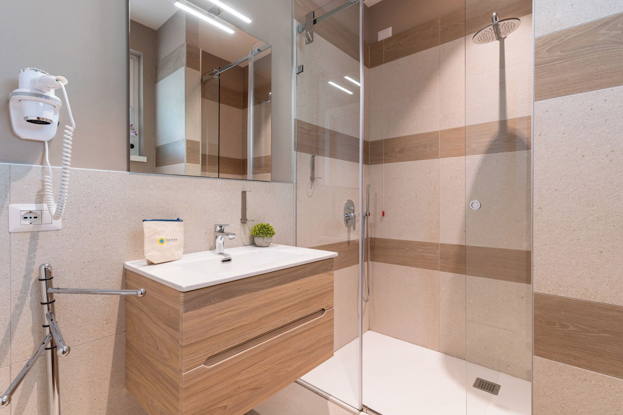 Een moderne badkamer met een douchecabine van glas, een houten wastafelmeubel met een wastafel en een haardroger bevestigd aan de muur.