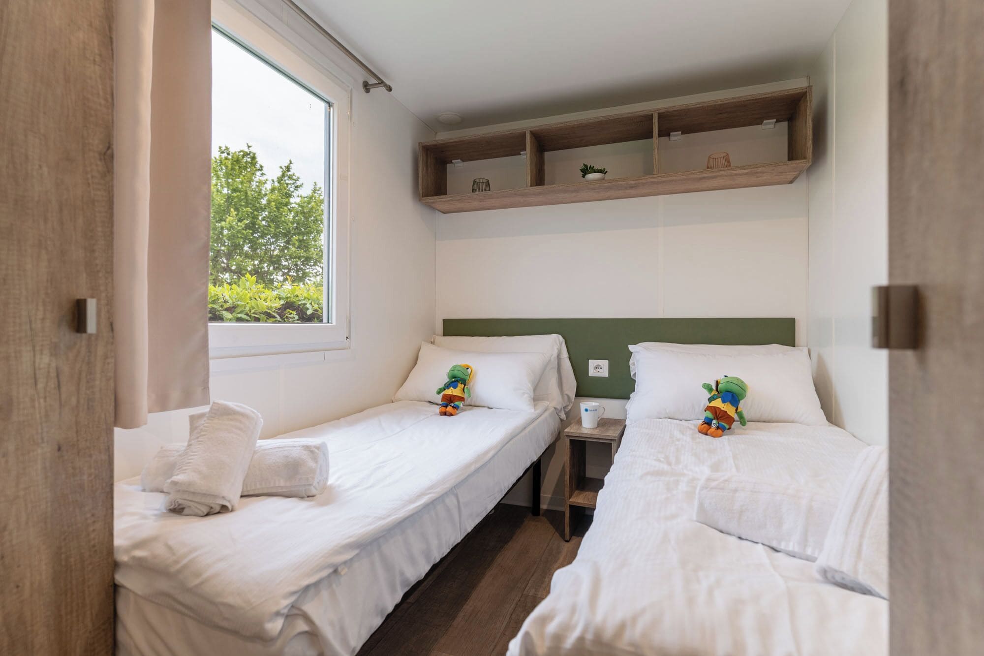 A room with two single beds, a small table, a shelf above the beds, and a window with a view of trees.