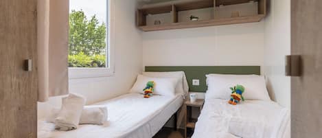 A room with two single beds, a small table, a shelf above the beds, and a window with a view of trees.
