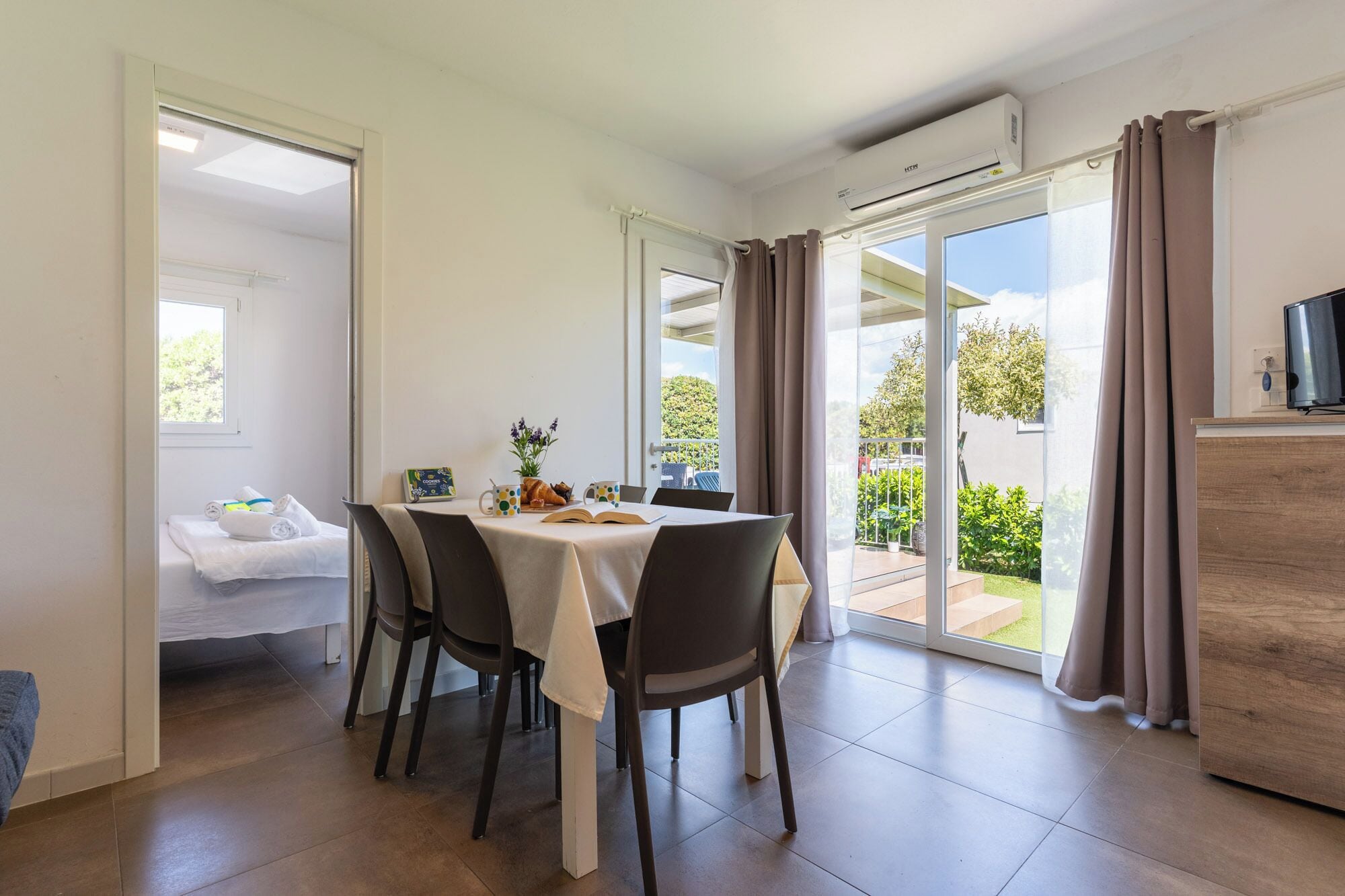 Un comedor con una mesa dispuesta para dos personas, una cama con edredón blanco y un balcón con vistas a la vegetación.