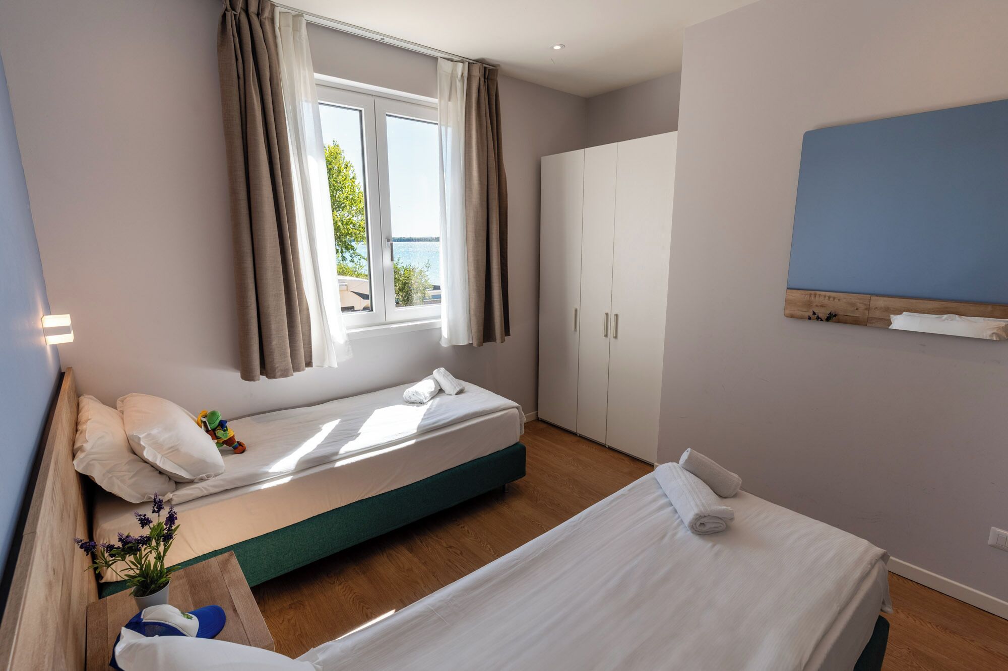 A bedroom with two beds, a wardrobe, a window with curtains, and a view of the outdoors.
