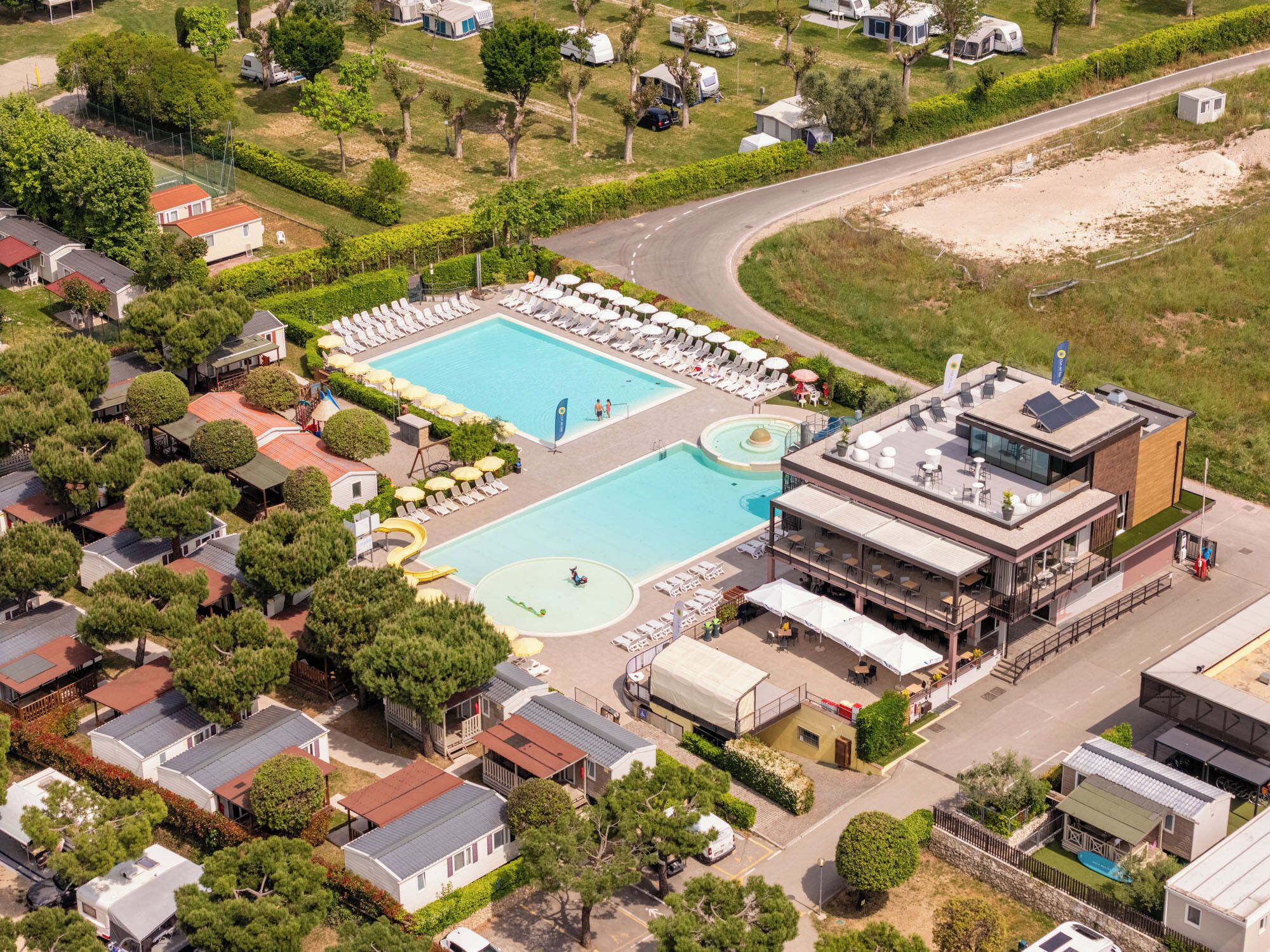 An aerial view of a resort with a large swimming pool, multiple lounge areas, and a variety of accommodation types including mobile homes and cabins.