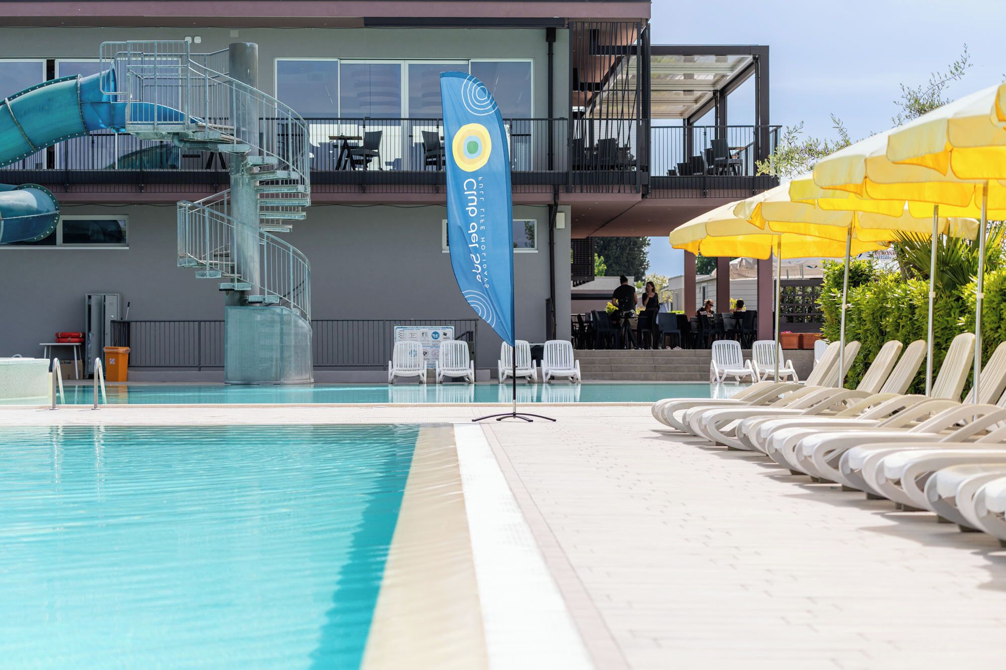 A swimming pool with lounge chairs, a water slide, and a building with outdoor seating.