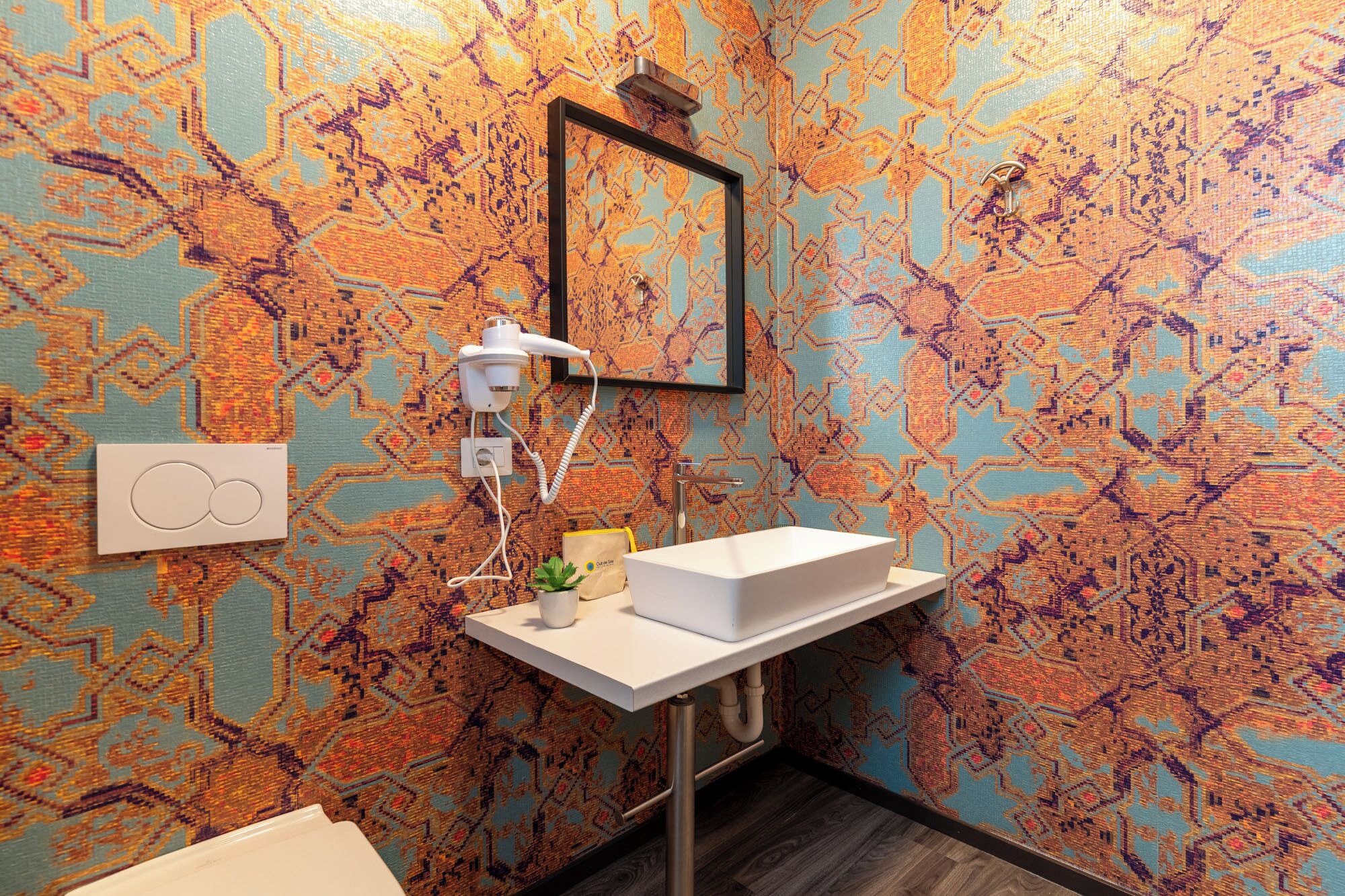 A bathroom with a patterned wall, a white sink, a hairdryer, and a small plant.