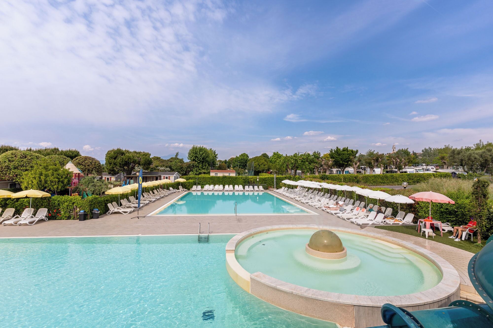 A swimming pool with lounge chairs and umbrellas, surrounded by greenery and buildings.