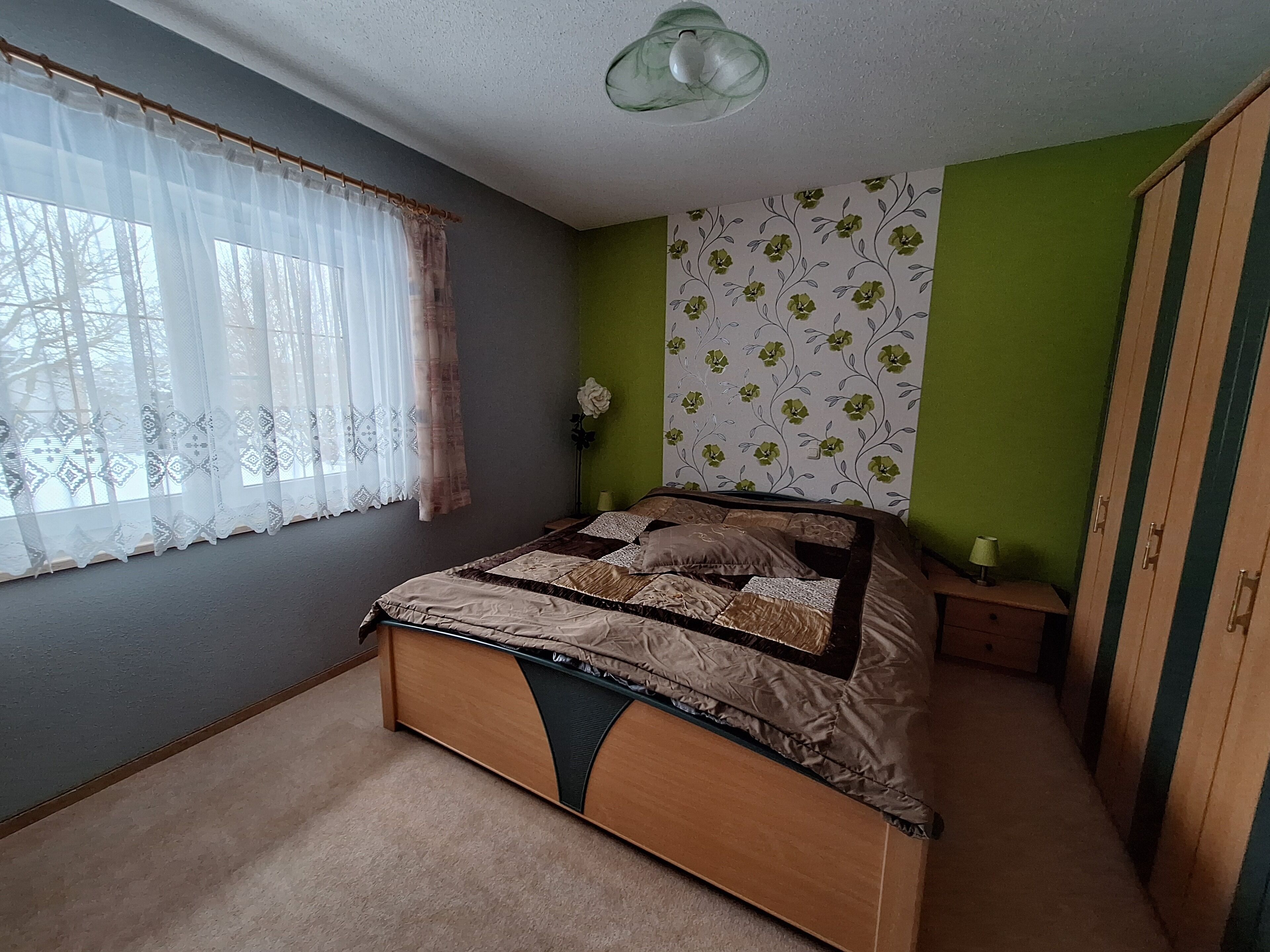 Ein Schlafzimmer mit einem Bett, einem Fenster mit durchsichtigen Vorhängen, einer grünen Wand mit Blumenwallpaper und einem Kleiderschrank.