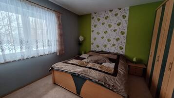 Ein Schlafzimmer mit einem Bett, einem Fenster mit durchsichtigen Vorhängen, einer grünen Wand mit Blumenwallpaper und einem Kleiderschrank.