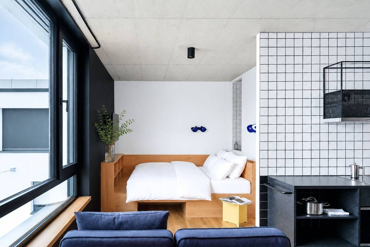 A modern hotel room with a bed, a small table, a plant, and a view through a large window.