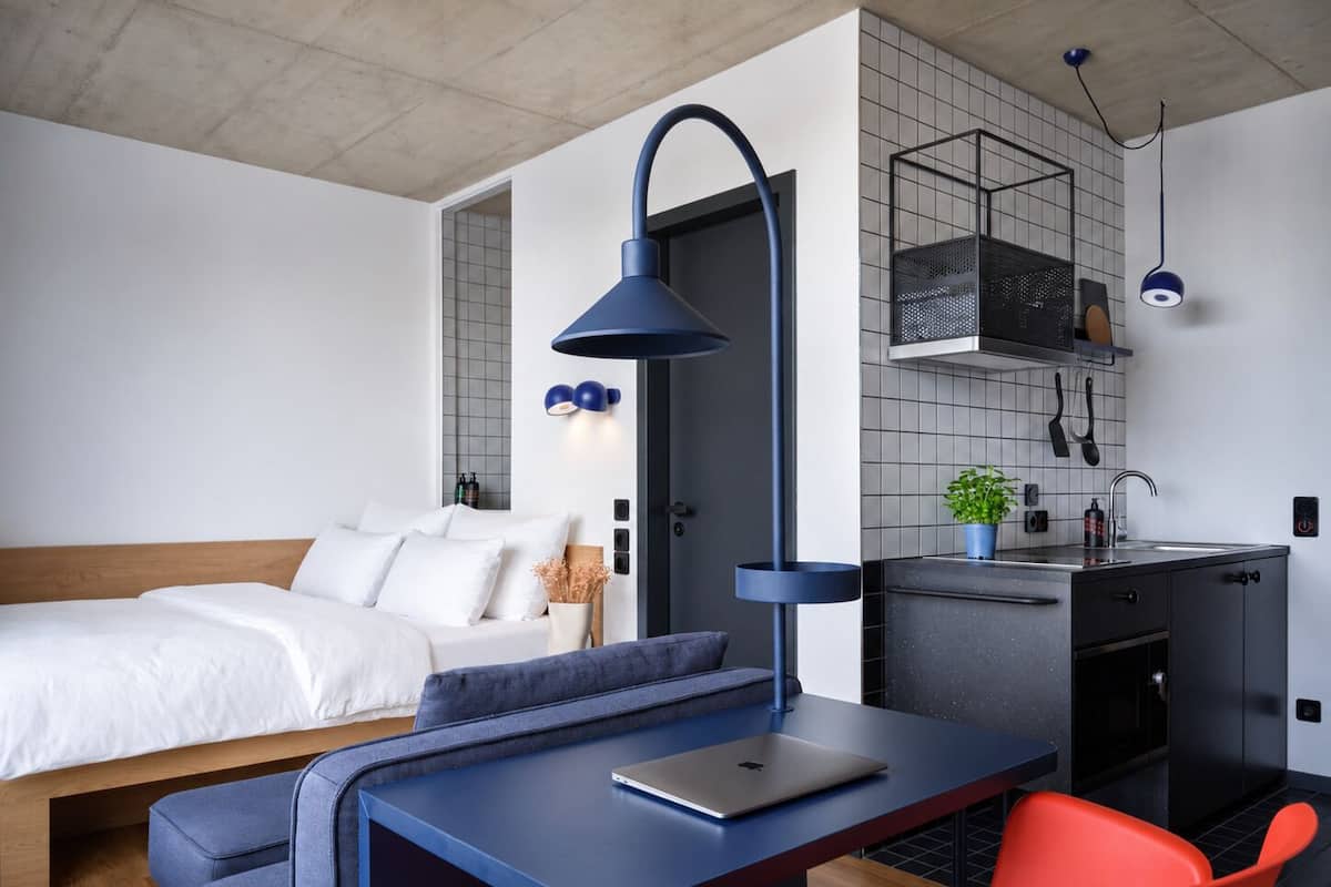 A modern hotel room with a bed, a desk with a laptop, a red chair, a blue lamp, and a kitchenette.