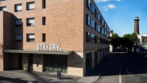 A modern building with a sign reading "STAYERY." It features a brick facade, multiple windows, and a clear sky with a few clouds.
