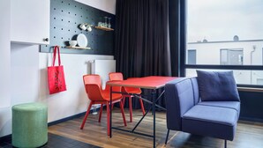 Ein modernes Zimmer mit einem roten Tisch, zwei roten Stühlen, einem blauen Sofa, einem grünen Hocker und einem Fenster mit Blick auf Gebäude.