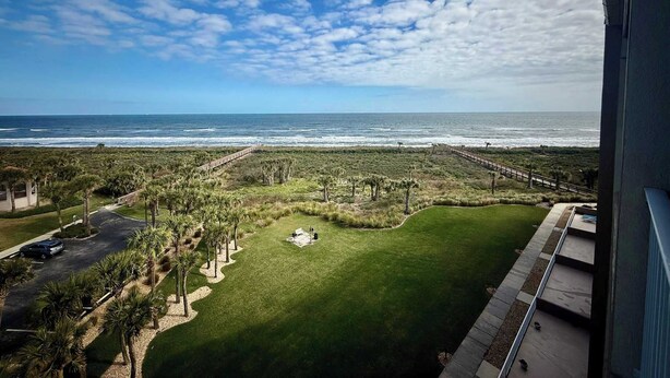 A view of a coastal landscape with a grassy area, palm trees, and a wooden boardwalk leading to the beach. - Rock the Sunset – Palm Coast Condo (Palm Coast)