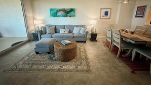 A living room with a grey sofa, a coffee table, a dining table with chairs, a chandelier, and a painting on the wall. - Rock the Sunset – Palm Coast Condo (Palm Coast)