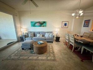 Ein Wohnzimmer mit grauem Sofa, einem Couchtisch, einem Esstisch mit Stühlen, einer Chandelierleuchtre und einem Bild an der Wand.