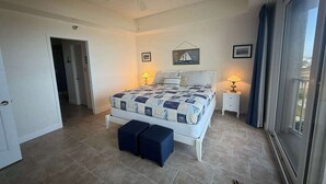 A bedroom with a bed, two bedside tables with lamps, a ceiling fan, a door, and a view through a window. - Rock the Sunset – Palm Coast Condo (Palm Coast)