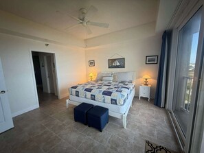 Ein Schlafzimmer mit einem Bett, zwei Nachttischen mit Lampen, einem Deckenventilator, einer Tür und Blick durch ein Fenster.