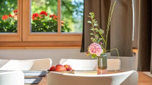 Une table à manger ornée d’un vase de fleurs, d’une assiette de fruits et offrant une vue sur un jardin par la fenêtre.