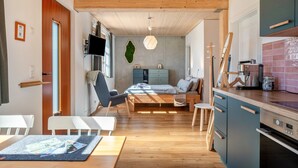 Un intérieur moderne avec un plafond en bois, un lit doté d’une tête de lit bleue, un coin repas avec une table et des chaises, une cuisine équipée d’une cuisinière et de placards, et un téléviseur fixé au mur.