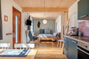 A modern interior with a wooden ceiling, a bed with a blue headboard, a dining area with a table and chairs, a kitchen with a stove and cabinets, and a wall-mounted TV. - Schwarzwaldgrün Holiday Apartment (Bonndorf im Schwarzwald)