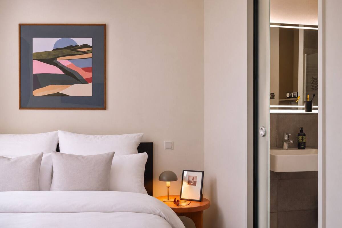A neatly made bed with white bedding, a framed abstract painting on the wall, a bedside table with a lamp and a framed photo, and a bathroom visible through an open door.