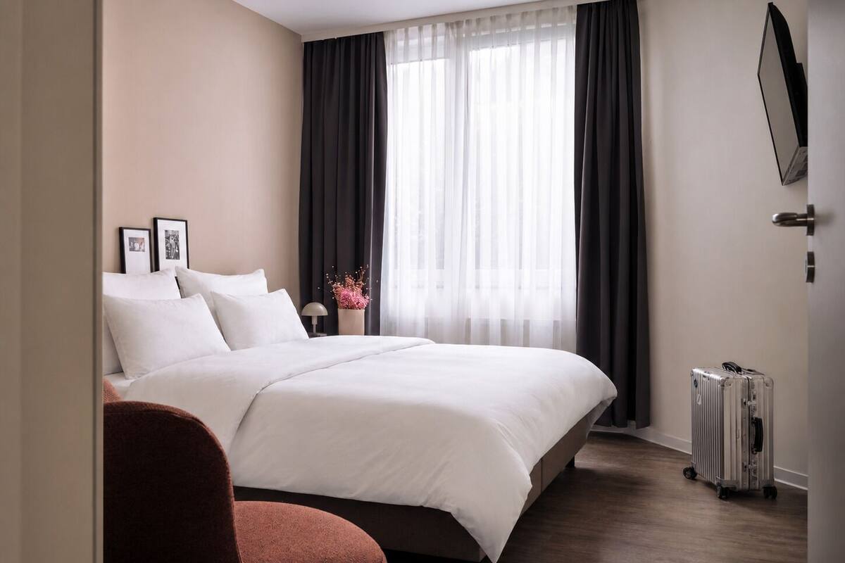 A hotel room with a large bed, a chair, a bedside table with a lamp, a mirror, and a suitcase.