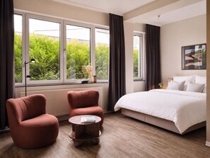 A modern hotel room with a large bed, two armchairs, a small round table, and a view of greenery through large windows. - STAYERY Cologne Sülz (Cologne)