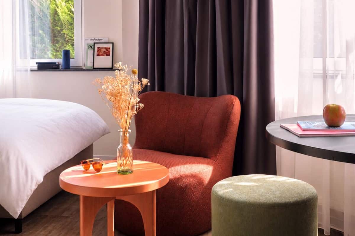 A hotel room with a bed, a red armchair, a small round table with a vase and glasses, and a green ottoman.