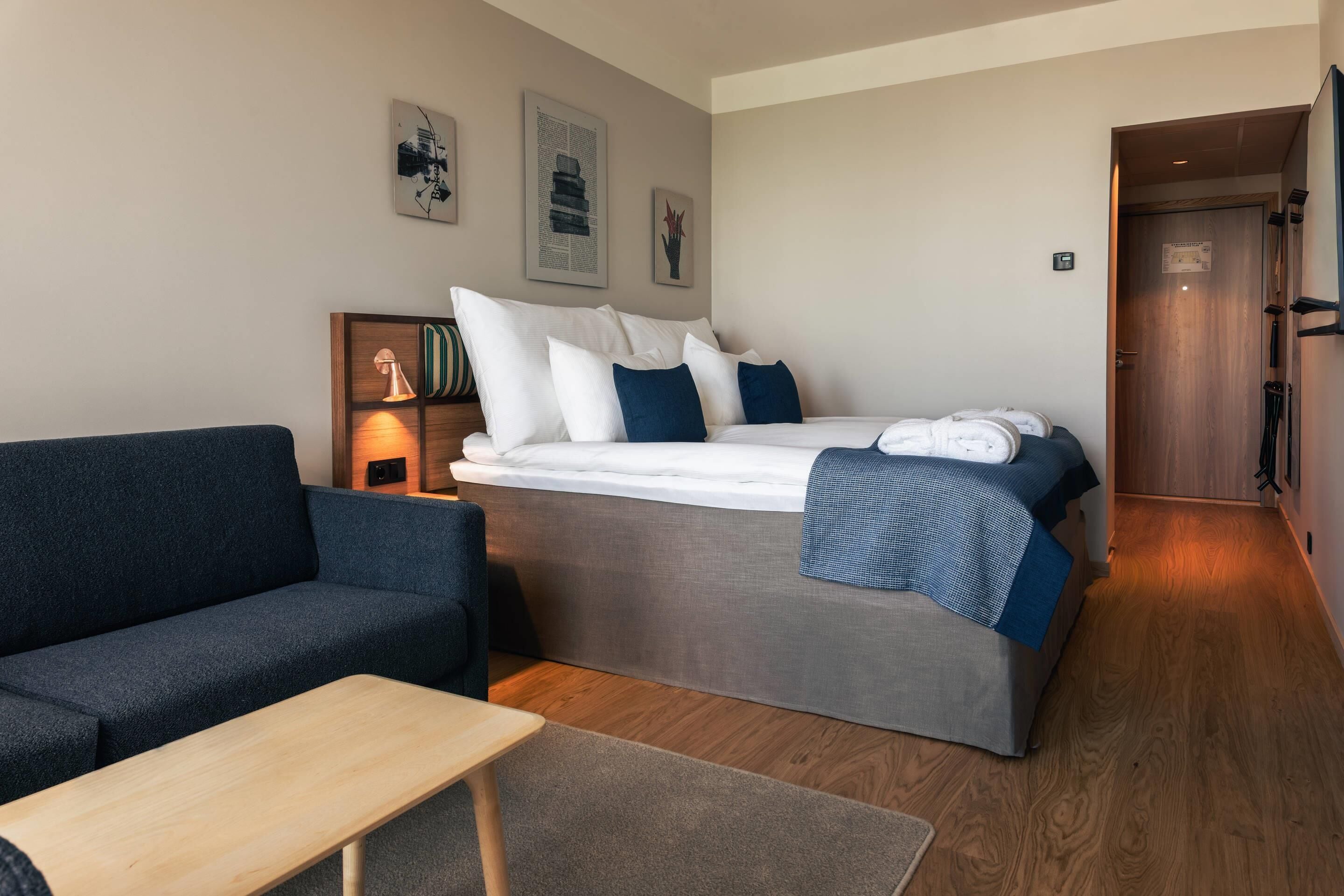 A modern hotel room with a bed, a sofa, a small table, and artwork on the walls.