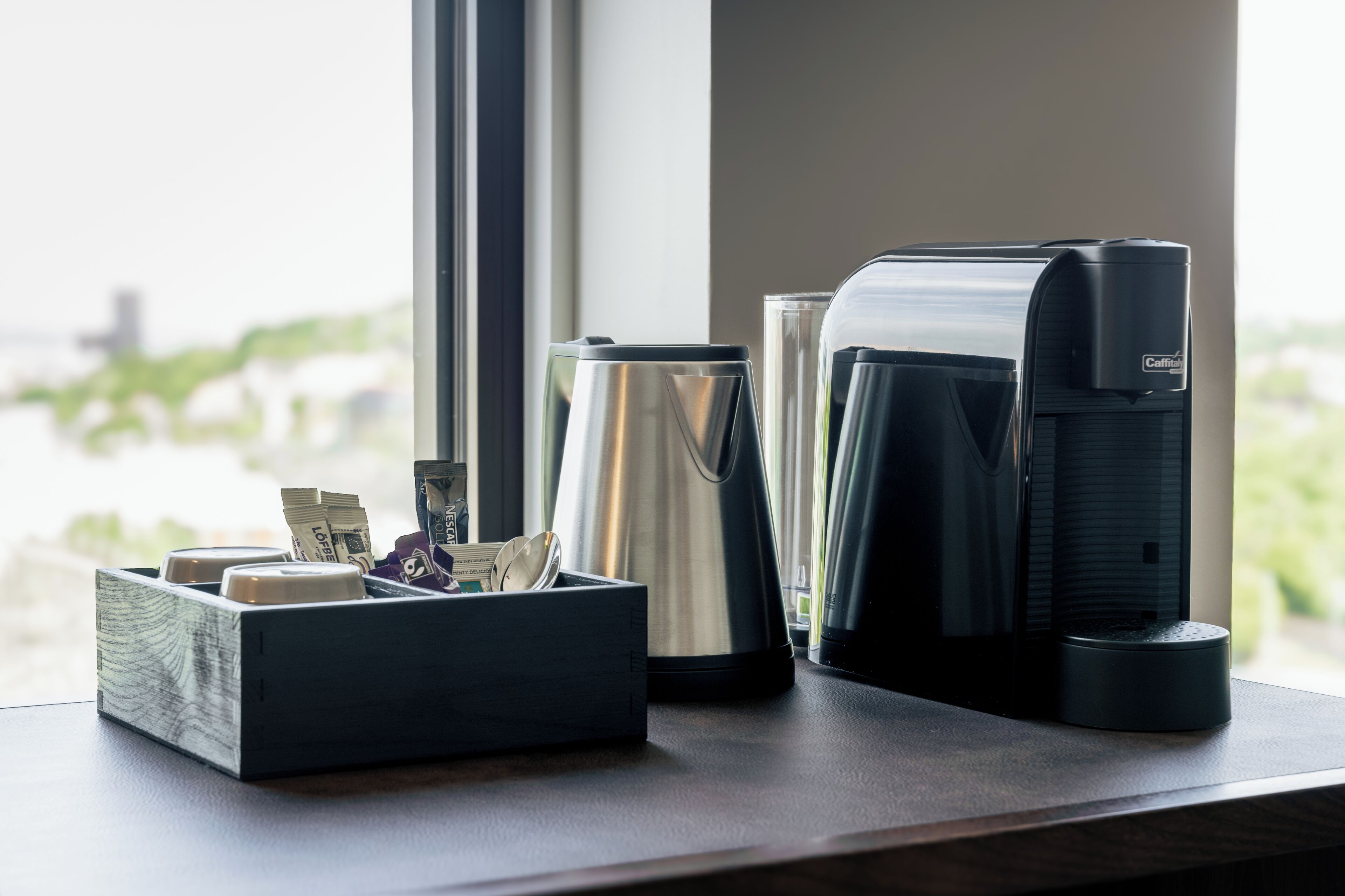 A coffee maker, a stainless steel kettle, and a wooden box with money and a card on a table by a window.