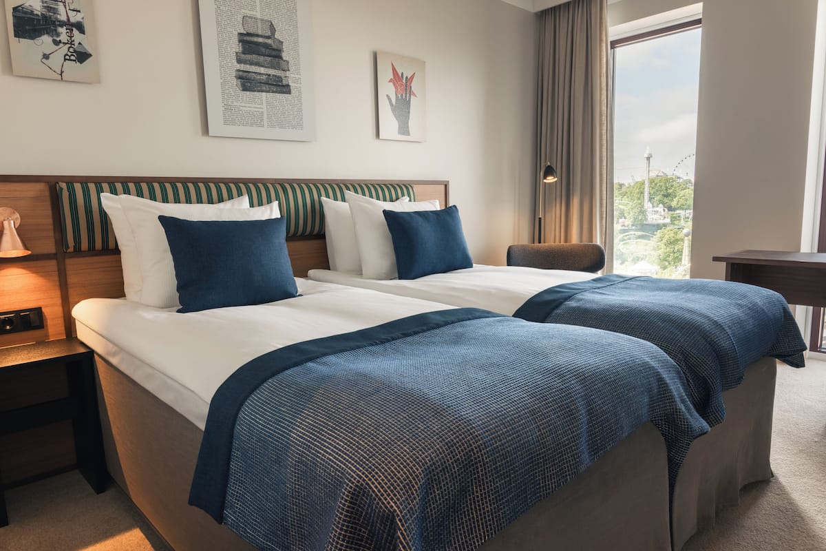 A hotel room with two beds, a nightstand, a lamp, and a view of a cityscape.