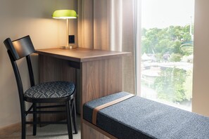 A room with a desk, a chair, a bench, a lamp, and a window with a view of buildings and trees. - Quality Hotel The Book (Gothenburg)