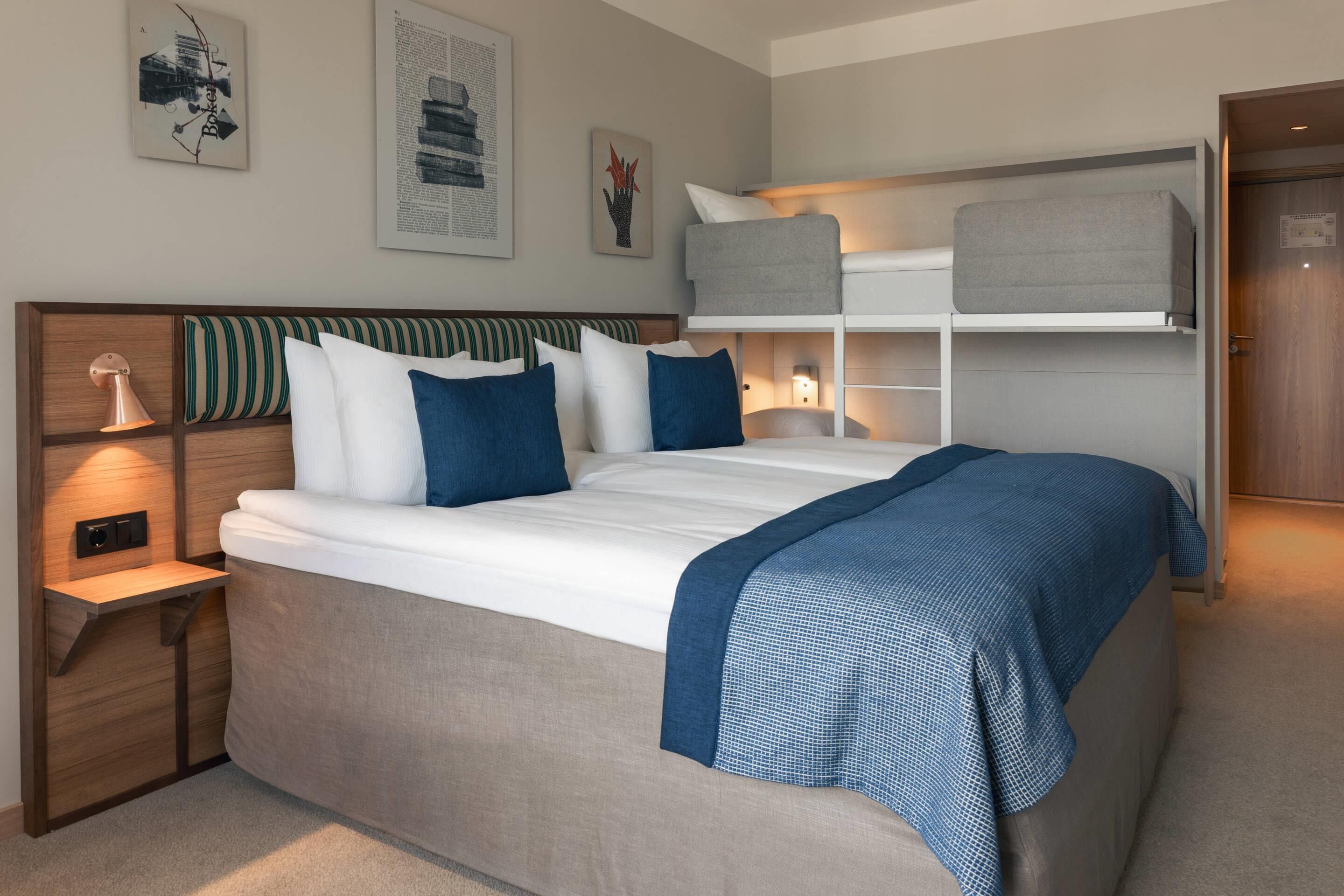 A modern hotel room with a large bed, a bedside table with a lamp, a built-in shelf with storage, and a bunk bed above.
