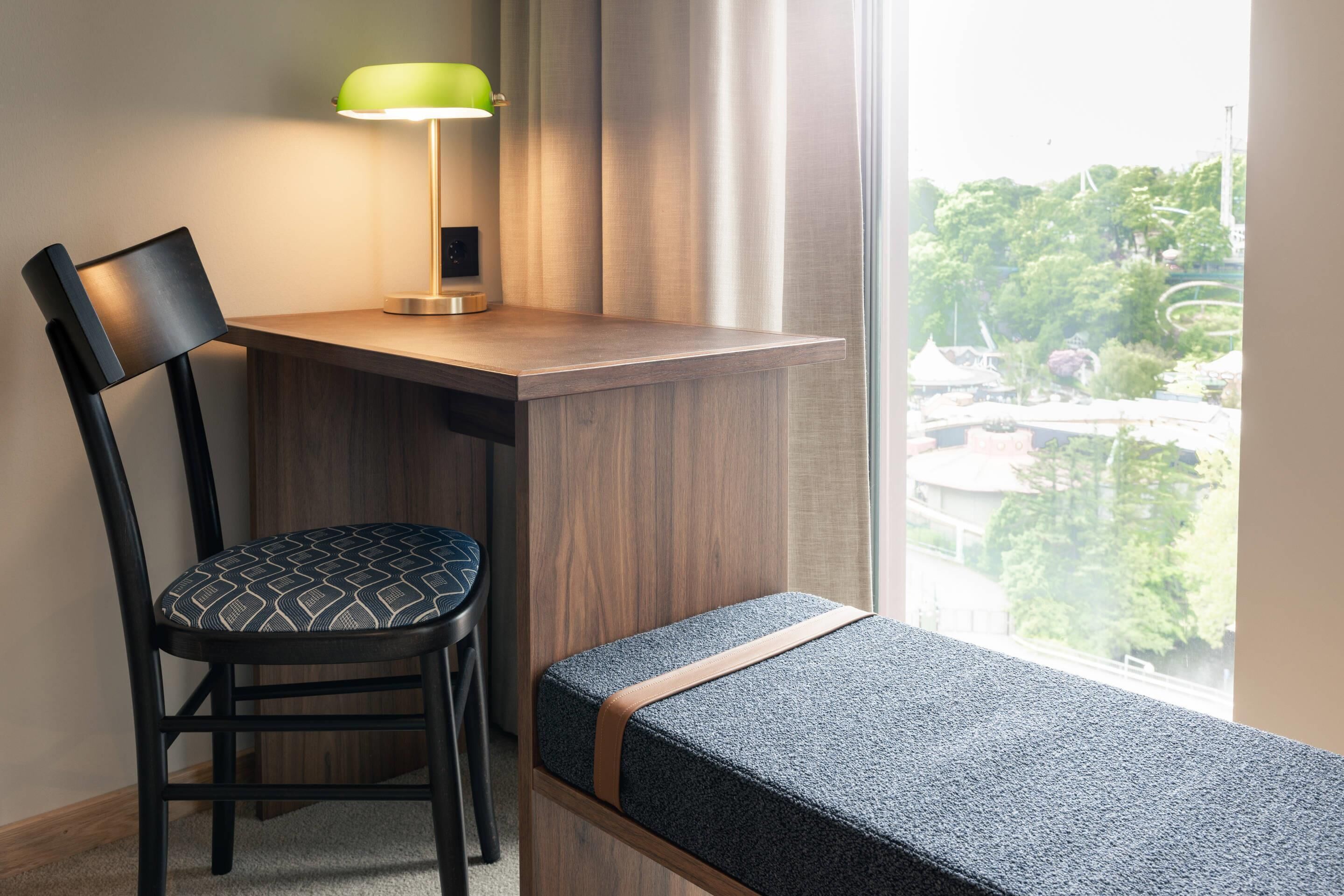 A room with a desk, chair, bench, lamp, and a view of greenery outside.