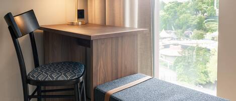 A room with a desk, chair, bench, lamp, and a view of greenery outside.