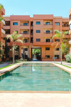 Un edificio moderno con una piscina central, rodeado de palmeras y un cielo despejado.