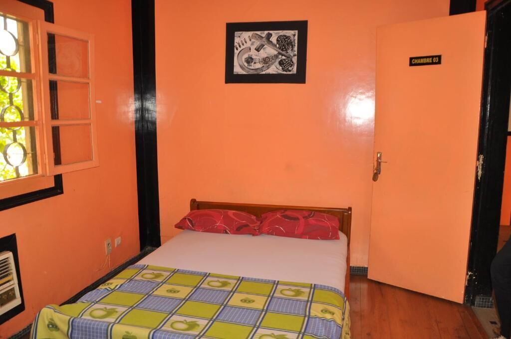Une pièce avec un mur orange, un lit recouvert d’une courtepointe à carreaux, une fenêtre donnant sur un paysage et une porte portant l’inscription « CHAMBRE 03 ».