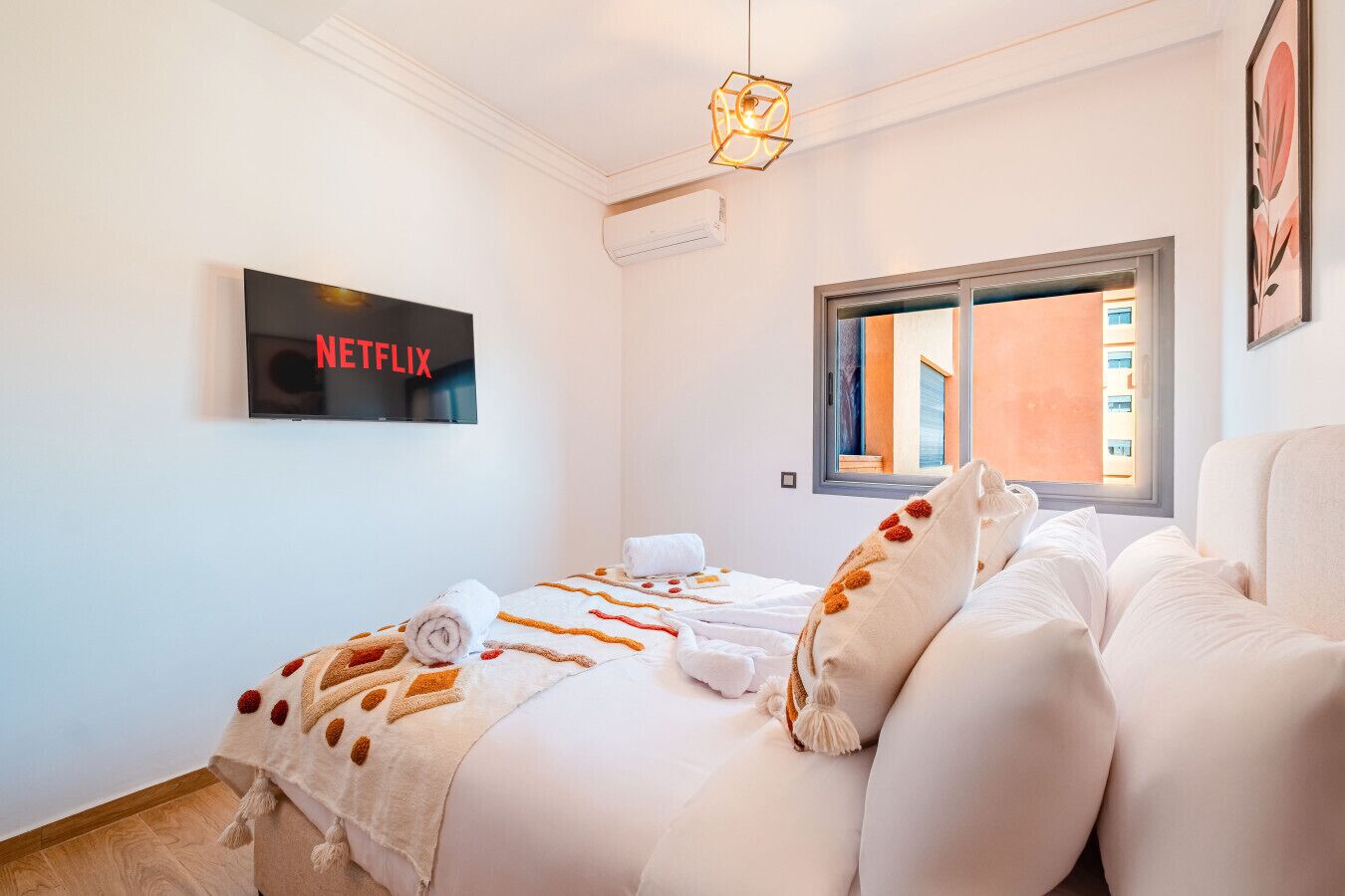 A hotel room with a bed, a TV mounted on the wall, a window with a view of buildings, and a ceiling light fixture.
