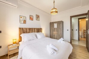 Una cama bien hecha con sábanas y toallas blancas, un cabecero de madera y una lámpara de noche. Hay tres cuadros enmarcados en la pared sobre la cama.