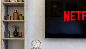 A living room with a large flat-screen TV mounted on the wall, wooden shelves with various decorative items, and a geometric light fixture on a console table. - Guéliz 2BR Near M Avenue – Piscine, Balcons & Clim (Marrakesh)