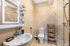Un baño con lavamanos, inodoro y cabina de ducha de vidrio. Hay repisas con plantas y artículos de aseo.
